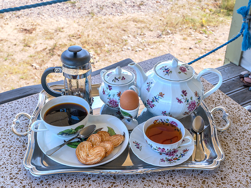 Plateau de petit déjeuner élégant complet avec viennoiseries, café, thé et vaisselle fine, posé sur une table de terrasse ensoleillée au bord de la mer.
