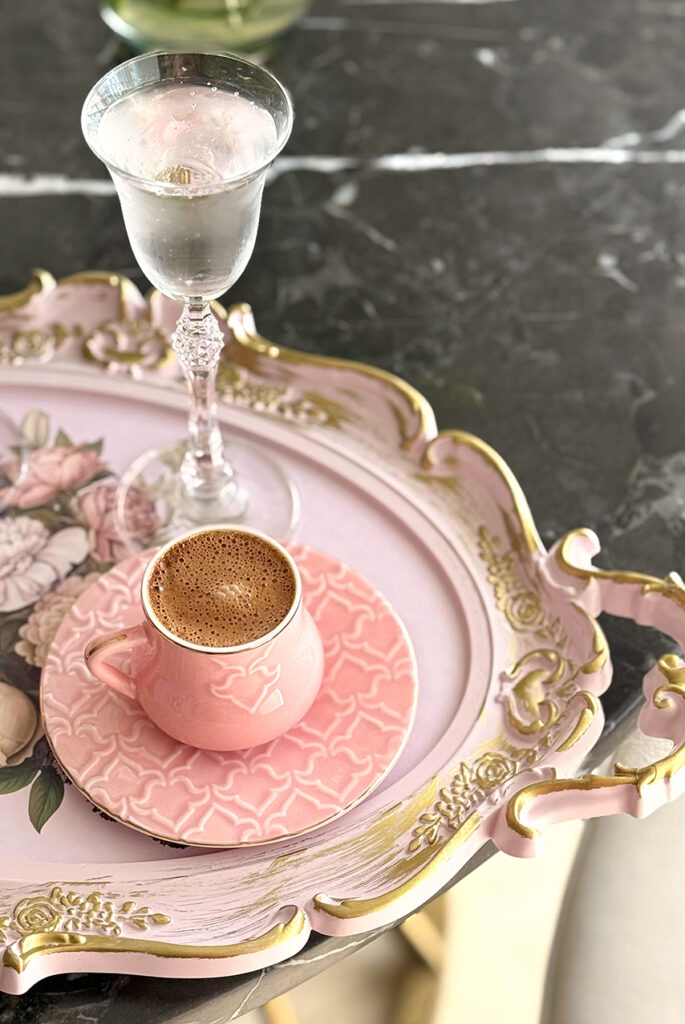 Plateau baroque rosé à dorures portant une tasse à café et soucoupe roses et un verre à pied en cristal, sur fond de marbre gris