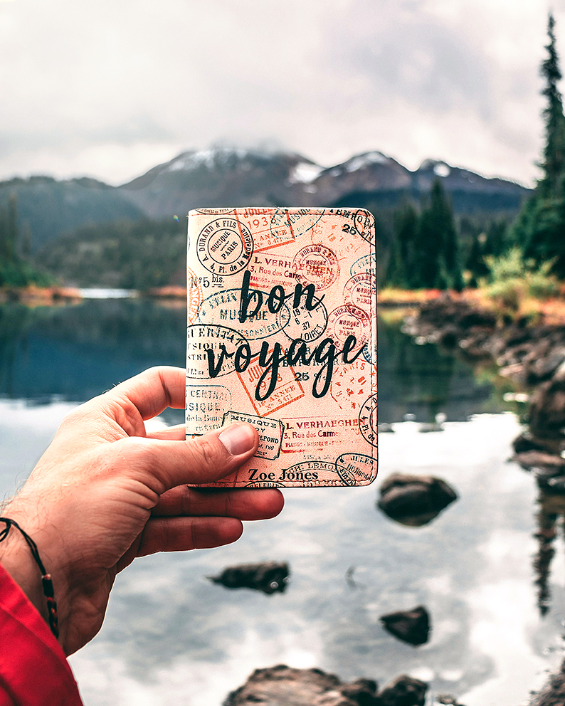 Une main tend un passeport couvert de tampons avec l'inscription "Bon voyage" sur fond de paysage montagneux aux cimes enneigées avec un lac bordé de sapins au premier plan